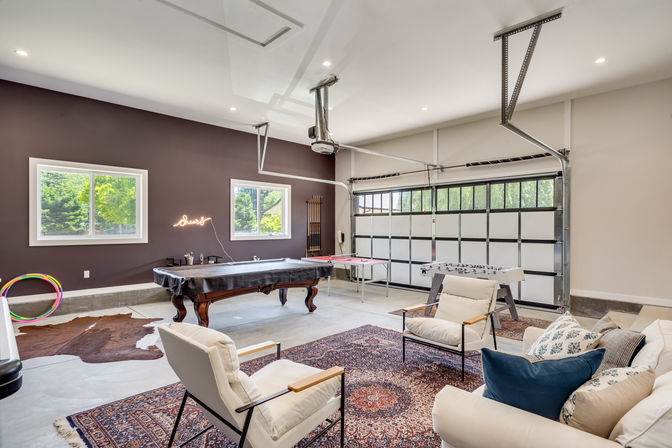 Bright converted garage game room with covered pool table, foosball and ping-pong tables, comfy lounge seating on layered rugs, neon wall sign, and a large glass-paneled garage door bringing in natural light and greenery.