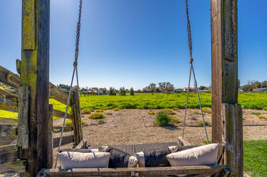 Inviting rustic wooden porch swing with cushions hanging from chains, overlooking a sunny green meadow and gravel path with distant houses beneath a clear blue sky.