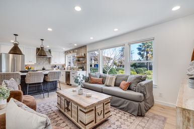 Sunlit open-concept living room and kitchen with gray slipcovered sofa, rustic trunk coffee table, rattan bar stools at the island, brass pendant lights, and large windows overlooking a landscaped garden.