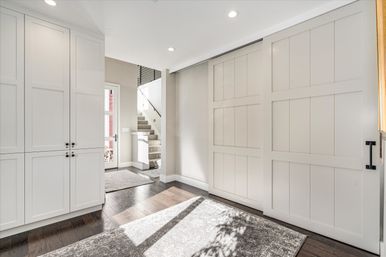 Sunlit modern foyer with white sliding barn doors, built-in cabinets, dark hardwood floors, area rug and carpeted staircase.