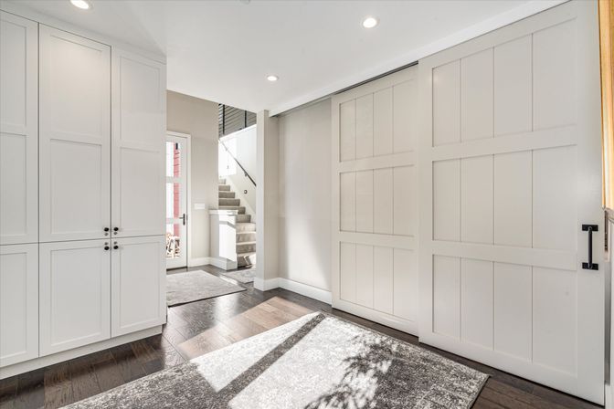 Sunlit modern foyer with white sliding barn doors, built-in cabinets, dark hardwood floors, area rug and carpeted staircase.