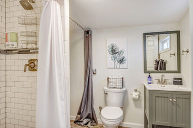 Bright, cozy home bathroom interior with subway-tile shower and white curtain, bronze shower fixtures, white toilet topped with a wire storage basket, gray vanity with mirror and sink, botanical wall art, and wood-look flooring.