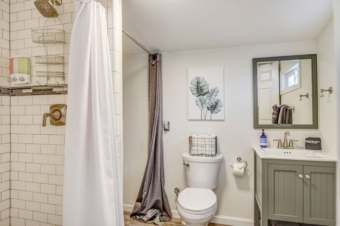 Bright, cozy home bathroom interior with subway-tile shower and white curtain, bronze shower fixtures, white toilet topped with a wire storage basket, gray vanity with mirror and sink, botanical wall art, and wood-look flooring.