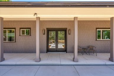 Covered front porch of a modern home with gray shingle siding, two square columns, double glass entry doors flanked by lantern sconces, concrete patio, and a small bistro table with two chairs.