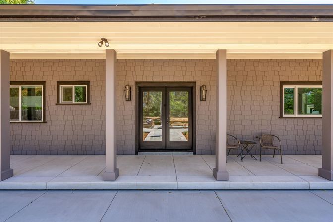 Covered front porch of a modern home with gray shingle siding, two square columns, double glass entry doors flanked by lantern sconces, concrete patio, and a small bistro table with two chairs.