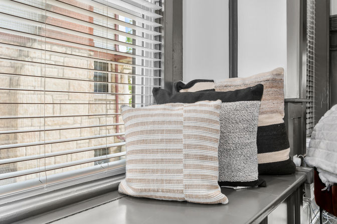 Cozy window seat with stacked beige, black and cream striped throw pillows on a gray windowsill next to white horizontal blinds and an urban exterior view.