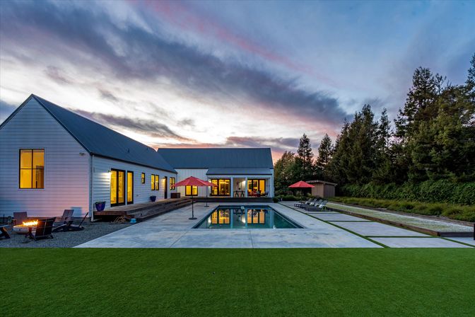 Modern white farmhouse-style home with metal roof overlooking a rectangular swimming pool at sunset, red umbrellas and lounge chairs on a concrete deck, cozy firepit seating to the left and manicured lawn with tall trees in the background.