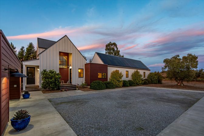Modern rural farmhouse with white board-and-batten siding and metal roofs at sunset, gravel driveway, potted plants and neatly trimmed shrubs under a pink-and-blue sky.