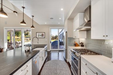 Bright open-concept modern kitchen with large dark island and farmhouse sink, pendant lights, stainless steel range and fridge, gray subway tile backsplash, hardwood floors and sliding glass doors opening to a sunny backyard deck.