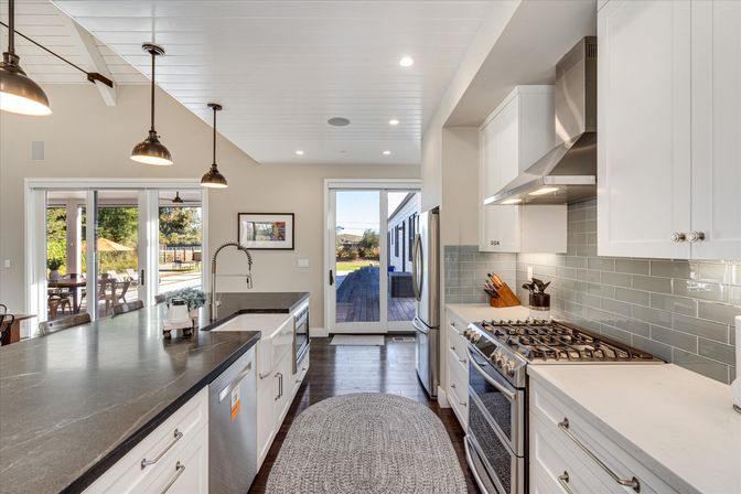 Bright open-concept modern kitchen with large dark island and farmhouse sink, pendant lights, stainless steel range and fridge, gray subway tile backsplash, hardwood floors and sliding glass doors opening to a sunny backyard deck.
