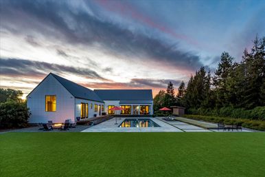 Modern white farmhouse at sunset with lit windows, rectangular reflecting pool, manicured lawn, patio seating, red umbrellas and tree-lined backyard.