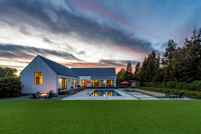 Modern white farmhouse at sunset with lit windows, rectangular reflecting pool, manicured lawn, patio seating, red umbrellas and tree-lined backyard.