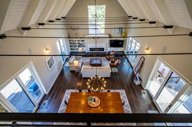 Aerial loft view of a sunlit open-concept living and dining room with vaulted white ceiling, dark hardwood floors, round chandelier, wooden dining table, cozy sofa, fireplace and sliding glass doors to a deck.