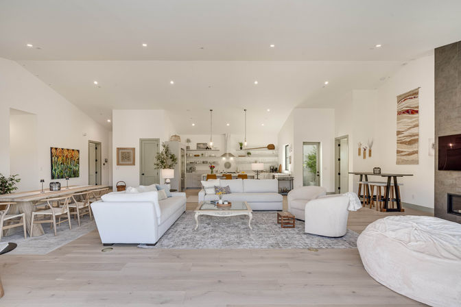 Bright, airy open-concept living room and kitchen in a modern home with vaulted ceiling, white sofas, round armchair, large area rug, long wooden dining table, pendant lights and neutral, cozy-yet-contemporary decor.