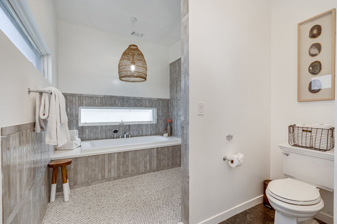 Bright modern master bathroom with built-in soaking tub in gray tile surround, narrow frosted window, woven rattan pendant light, pebble-mosaic floor, towel rack and wooden stool, and toilet with wire basket — spa-like neutral design.