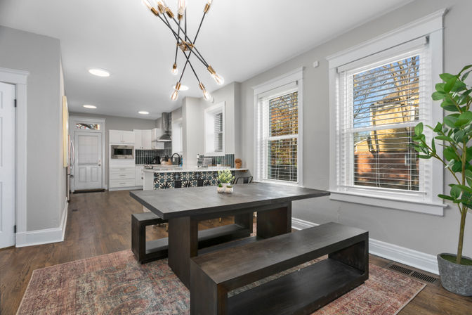 Light-filled open-plan dining and kitchen with dark wood bench table, patterned tile island, modern sputnik chandelier, hardwood floors, neutral gray walls, and large white-trim windows.
