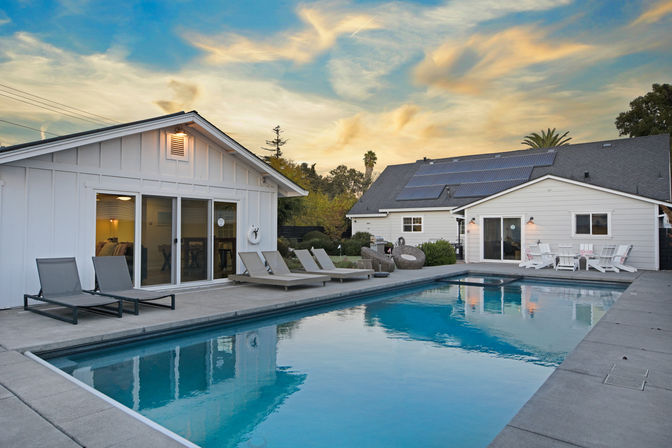 Sparkling backyard swimming pool at sunset beside a white modern suburban home with lounge chairs, wicker seating, Adirondack chairs and solar panels on the roof.