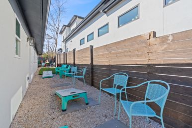 Narrow backyard seating area beside a white stucco building, featuring turquoise metal chairs, cornhole boards on a gravel path, a wooden privacy fence and small patch of artificial turf.