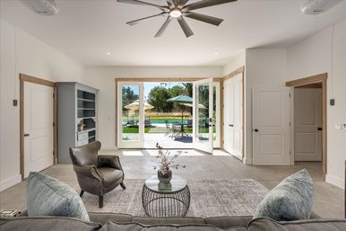 Bright modern open-plan living room with a large ceiling fan, gray sofa and leather armchair around a round coffee table, wide French doors opening to a sunny patio with umbrella-shaded dining set and a swimming pool