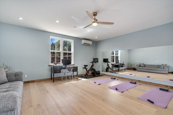Light-filled home gym and office with a mirrored wall, two stationary bikes with screens, three purple yoga mats on hardwood floors, gray sofa and desk under a window.