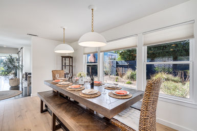 Sunlit open-concept dining room with rustic wooden table set for six, woven rattan chairs and bench, white pendant lights, and large windows overlooking a landscaped backyard garden.