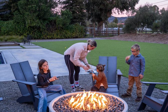 Adult and three children gathered around a backyard fire pit at dusk, roasting marshmallows on outdoor chairs by a green lawn with trees and a fenced yard in the background.