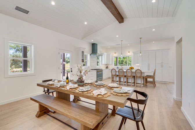 Bright open-concept modern farmhouse kitchen and dining area with a long wooden table and bench set, white cabinetry, large island with round-back stools, pendant lights, vaulted shiplap ceiling with exposed beam, and light hardwood floors