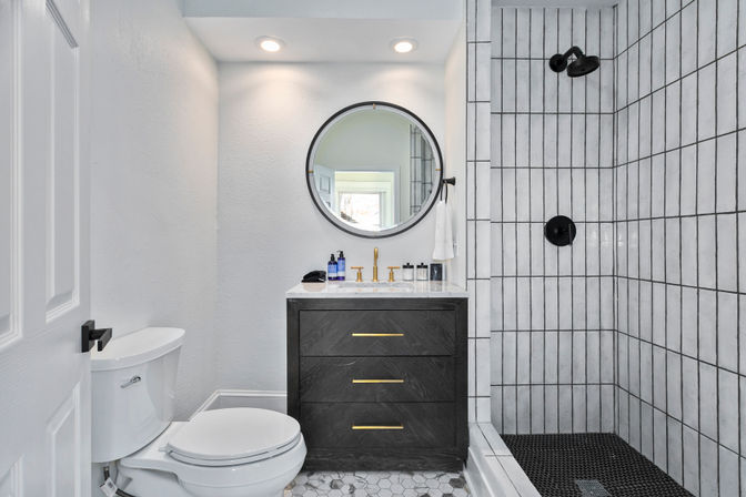 Stylish modern residential bathroom with black three-drawer vanity, marble countertop and brass faucet under a round mirror, white toilet, and walk-in shower with vertical white tiles and matte black fixtures on hexagon floor tiles.