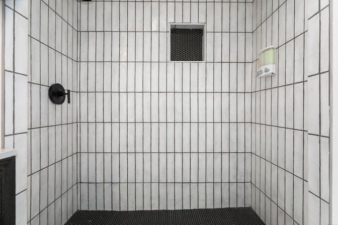 Modern walk-in shower with vertical white tiles and dark grout, black hexagon floor tiles, recessed niche, wall soap dispenser, and matte-black shower handle