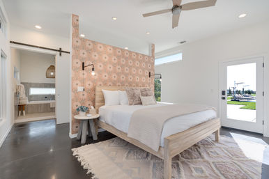 Sunlit modern bedroom with a wooden platform bed against a pink floral partition wall, wall-mounted lamps, textured throw pillows and rug, sliding barn door to a bright ensuite, and a glass door opening to a backyard pool patio.