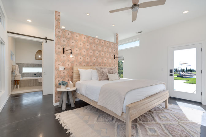 Sunlit modern bedroom with a wooden platform bed against a pink floral partition wall, wall-mounted lamps, textured throw pillows and rug, sliding barn door to a bright ensuite, and a glass door opening to a backyard pool patio.