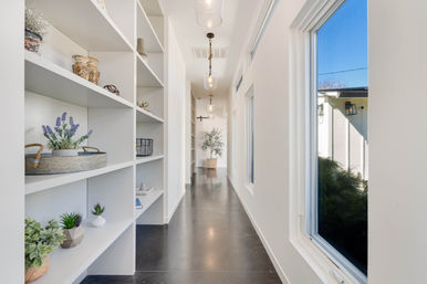 Sunlit modern hallway in a contemporary home with built-in white shelving styled with plants and baskets, polished dark concrete floor, hanging pendant lights, and large windows letting in natural light.