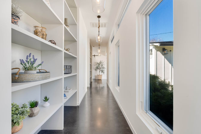 Sunlit modern hallway in a contemporary home with built-in white shelving styled with plants and baskets, polished dark concrete floor, hanging pendant lights, and large windows letting in natural light.