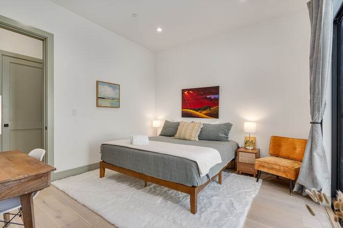 Cozy minimalist modern bedroom interior with a wooden platform bed in gray linens, fluffy white rug, bedside lamps, orange velvet accent chair and large window with curtains.