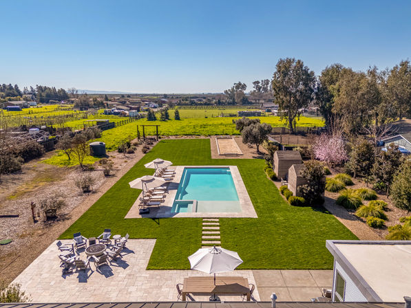 Aerial view of a rectangular backyard pool on a sunny manicured lawn, with lounge chairs and umbrellas, stone patio with fire pit and outdoor dining, set against open countryside.