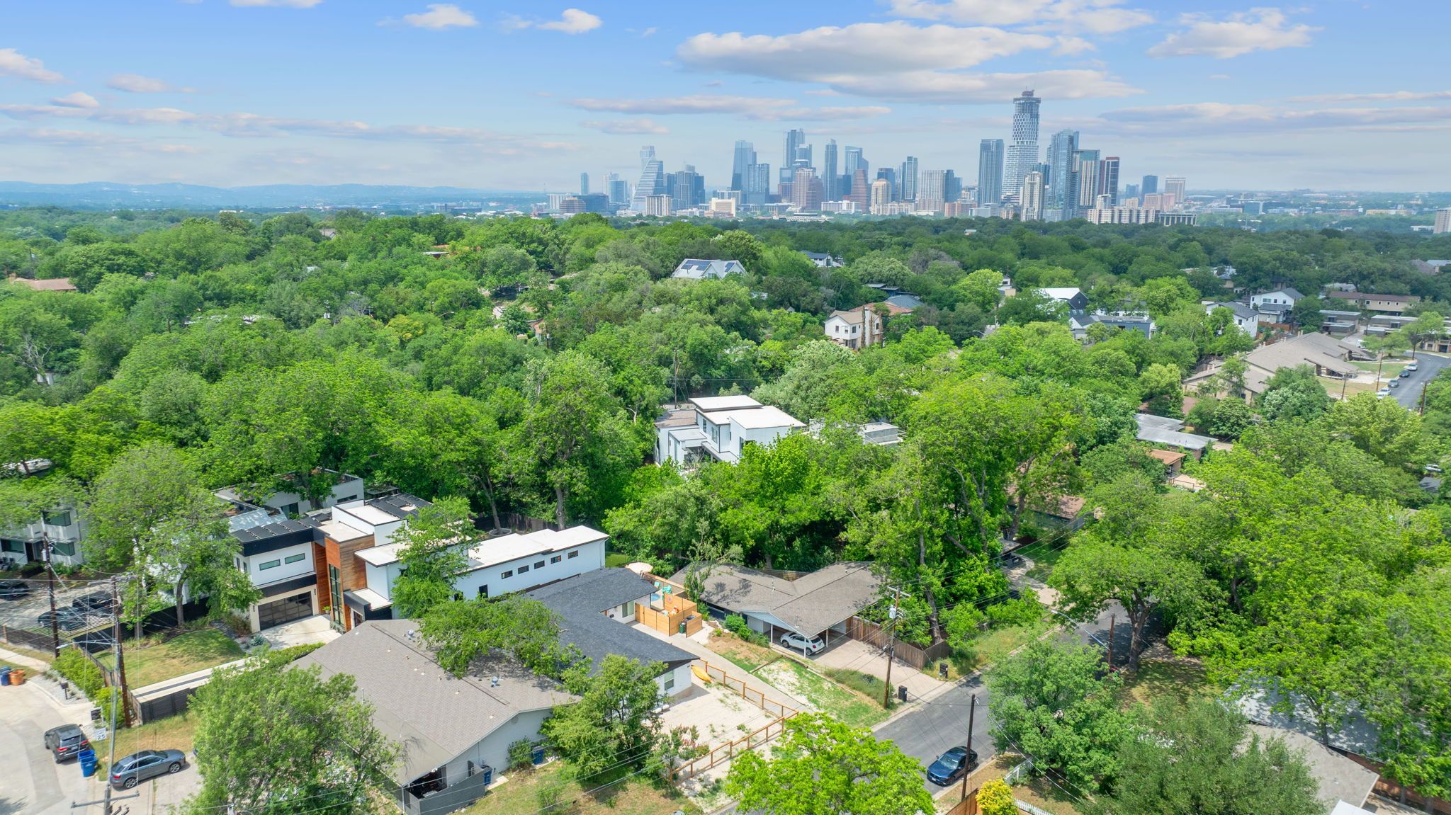 Aerial view of a lush, tree-lined residential neighborhood with modern houses in the foreground and a distant downtown skyline under a blue sky.