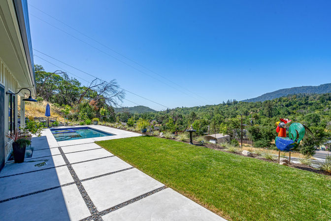 Modern backyard with concrete paver patio and rectangular plunge pool, manicured lawn, colorful metal rooster sculpture, and panoramic tree-covered hills under a clear blue sky.