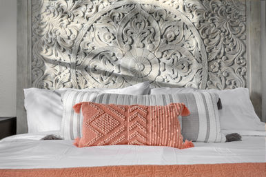Boho-chic bedroom with carved whitewashed floral mandala headboard, white and gray striped pillows, coral textured lumbar pillow with tassels, and white bedding topped by a coral quilt.