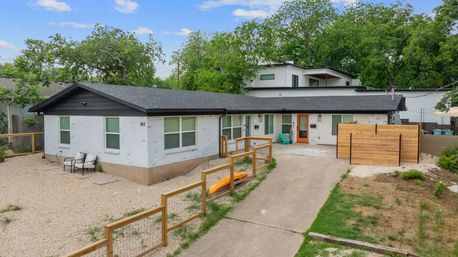 Single-story white brick ranch house with black roof and orange front door, gravel front yard with wooden fence, driveway and an orange kayak, leafy suburban trees in background.