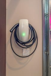 Tesla wall-mounted EV charger in a home garage on a beige wall, coiled black charging cable looped around it with a glowing green status light.