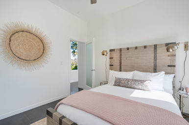 Bright coastal-style bedroom with a rustic wood headboard, crisp white bedding and pink quilt, decorative bolster pillow, brass wall sconces, round woven wall art, and an open door showing a palm-tree view.