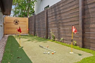 Backyard putting green on artificial turf with three red-flag holes, crossed putters and golf balls, dark wooden privacy fence and light wood gate with decorative star.