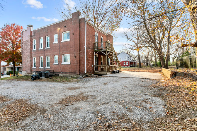 Two-story red brick residential building with wooden exterior staircase and porch, gravel parking area and concrete pad, surrounded by autumn trees with fallen leaves in a quiet neighborhood.