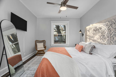 Cozy modern bedroom with carved wood headboard, white linens and coral throw, rattan accent chair, full-length mirror, wall-mounted TV, ceiling fan, and tree-view window.