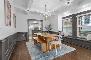 Sunlit Craftsman-style dining room with coffered ceiling and brass chandelier, long wooden farmhouse table and bench, white spindle chair, blue area rug, gray wainscoting, built-in window seat with pillows, hardwood floors, open to a white kitchen.