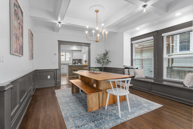 Sunlit Craftsman-style dining room with coffered ceiling and brass chandelier, long wooden farmhouse table and bench, white spindle chair, blue area rug, gray wainscoting, built-in window seat with pillows, hardwood floors, open to a white kitchen.