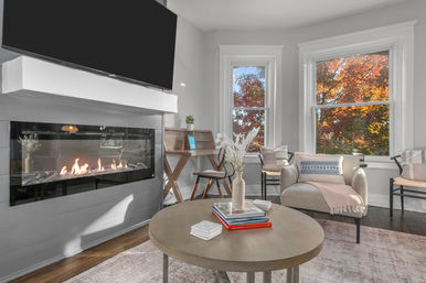 Cozy modern living room with wall-mounted TV and linear gas fireplace, round coffee table stacked with books and vase, neutral armchair with throw, and two tall windows framing autumn foliage