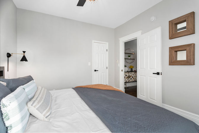 Bright modern bedroom interior with gray walls and white trim, bed dressed in blue-gray quilt and striped pillows, black swing-arm wall lamp, open white door showing hallway with round wall shelf and decorative console, and two wood-framed square mirrors on the wall.
