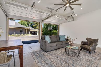 Bright converted garage living space with open double garage doors to a suburban driveway and greenery; gray sofa with teal pillows, leather armchair, round wire‑frame coffee table on a patterned rug, wooden desk and ceiling fan—modern indoor‑outdoor lounge.