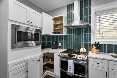 Sleek modern kitchen with white shaker cabinets, deep teal vertical subway-tile backsplash, stainless steel range and vent hood, built-in microwave, open corner cabinet with pots, coffee maker, knife block, and a window with blinds.